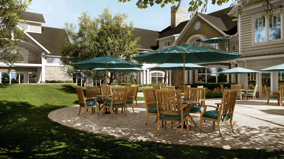 Outdoor patio area at Broadview Senior Living at Purchase College featuring multiple wooden tables with green cushioned chairs and large green umbrellas. The patio is surrounded by well-maintained grass, trees, and a large building with multiple windows in the background under a clear blue sky.