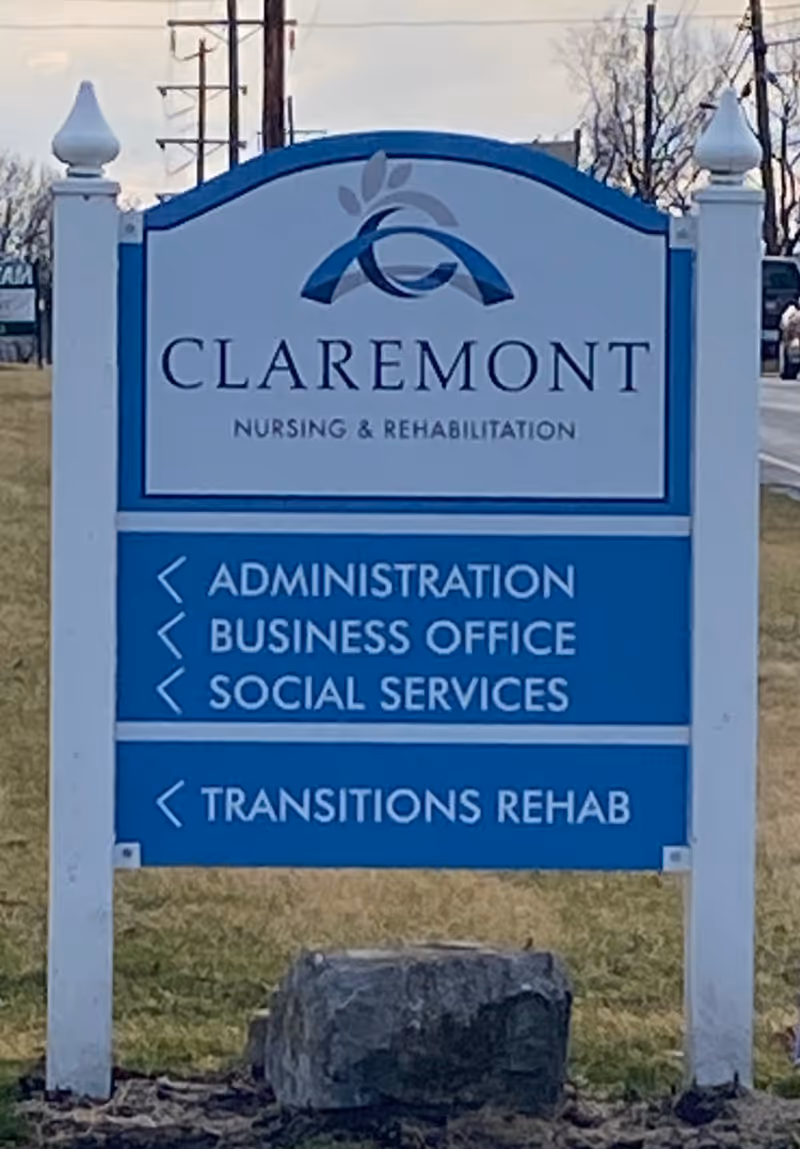 Outdoor sign for Claremont Nursing & Rehabilitation Center with directions to Administration, Business Office, Social Services, and Transitions Rehab. The sign is white and blue with a logo at the top and is situated on a grassy area near a road.