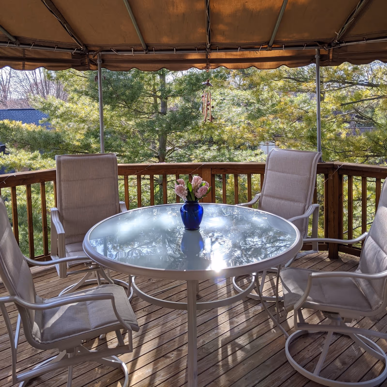 A covered outdoor wooden deck with a round glass table and four beige cushioned chairs. A blue vase with pink flowers is placed in the center of the table. Green trees are visible in the background.