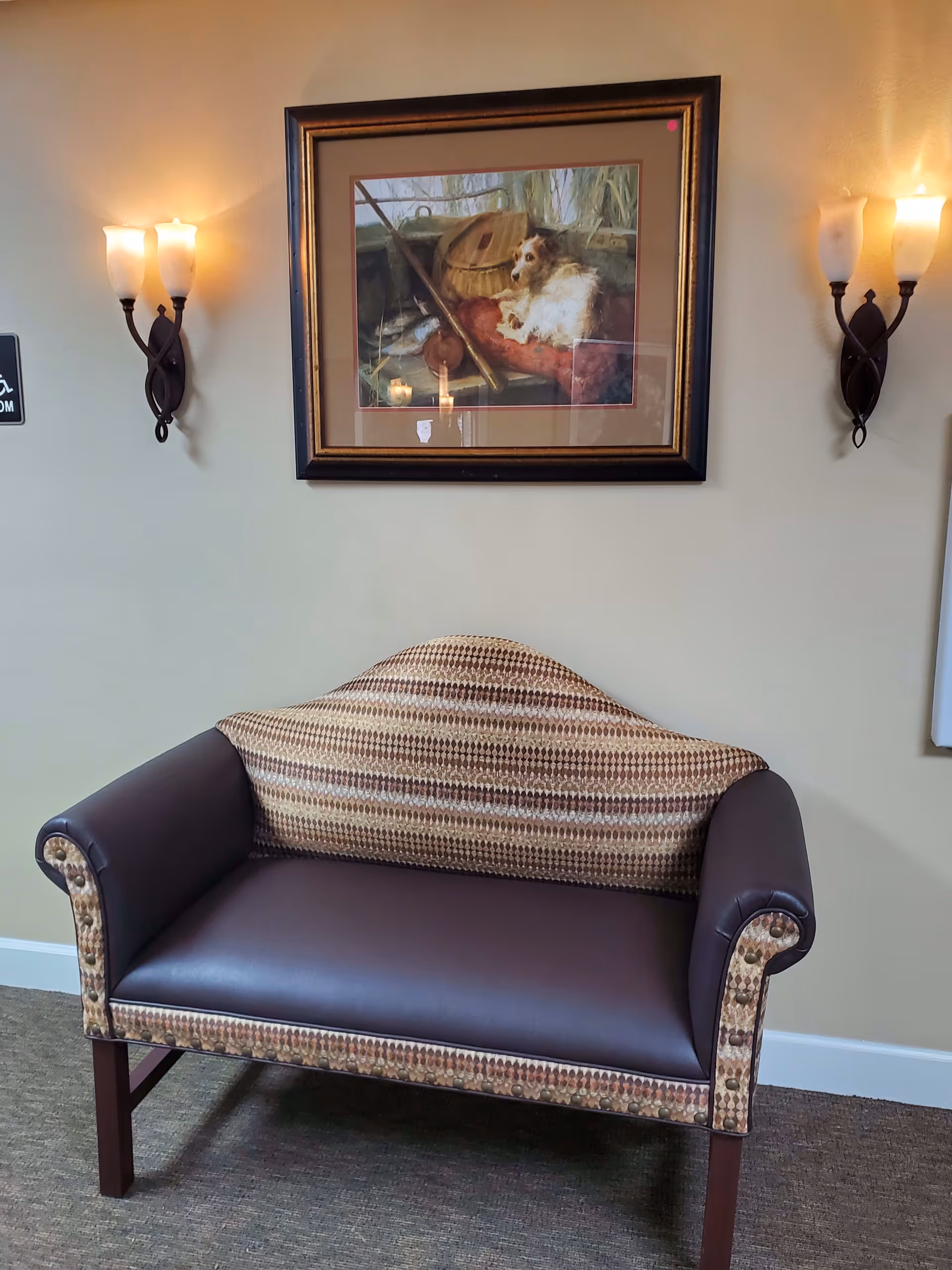 A small upholstered bench with patterned fabric and dark brown leather seat placed against a beige wall. Above the bench hangs a framed painting of a dog resting on a red cushion with fishing equipment in the background. Two wall-mounted candle-style light fixtures with frosted glass shades flank the painting.