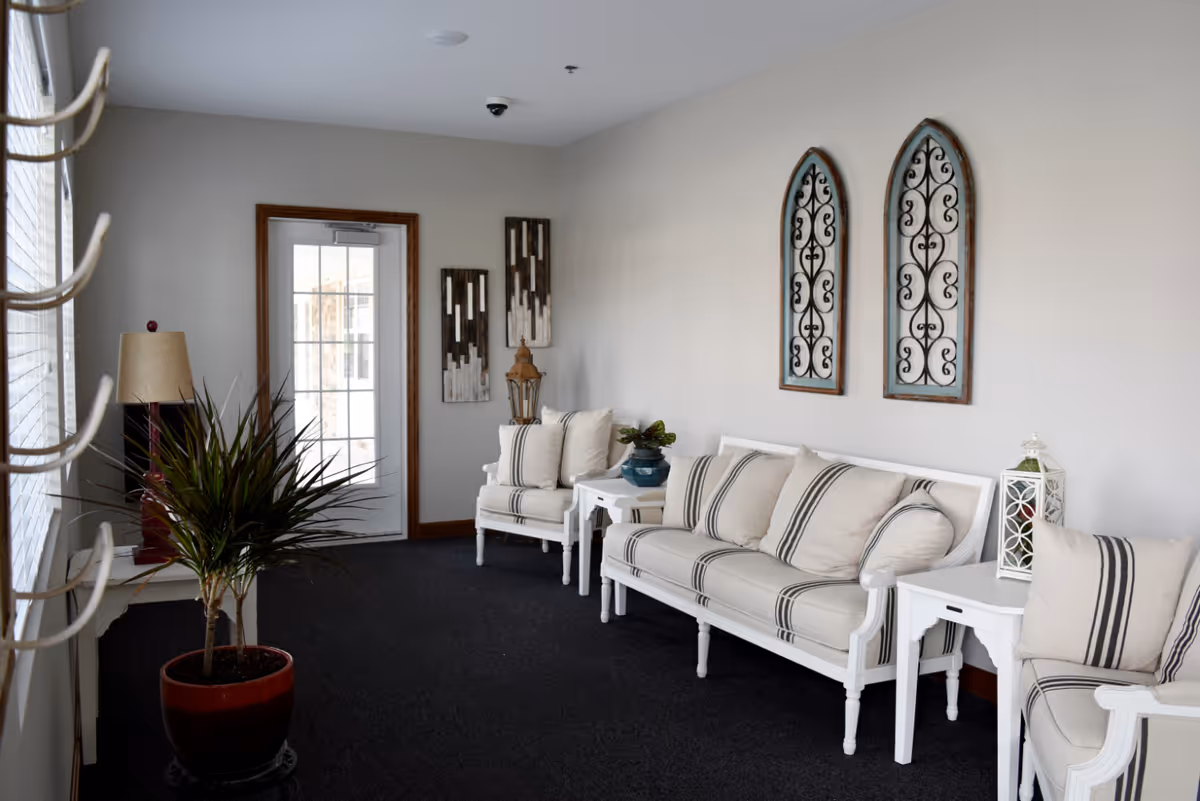 A bright and cozy sitting area with white cushioned chairs and a sofa featuring striped pillows. The room has a dark carpet, light gray walls, decorative wall art, a potted plant, a lamp, and a glass door with wooden trim leading outside.
