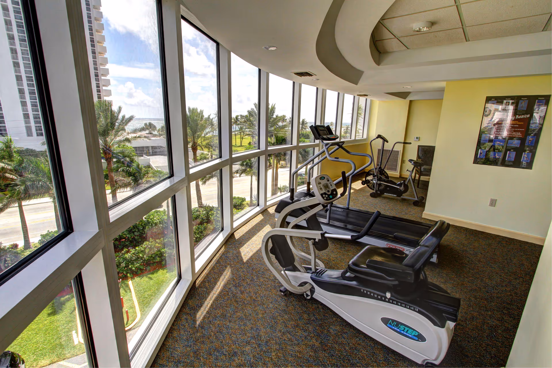 A fitness room with large curved floor-to-ceiling windows overlooking palm trees and a street outside. Inside the room, there are exercise machines including a recumbent bike, a treadmill, and an elliptical trainer. The walls are light yellow, and there is a framed poster on one wall.