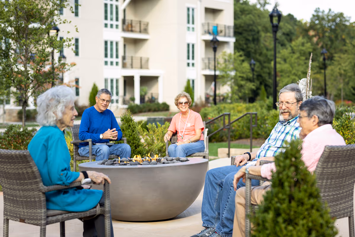 A group of five elderly people sitting around a circular outdoor fire pit in a garden area of a senior living facility. They are seated on wicker chairs, enjoying the fire and each other's company. In the background, there is a multi-story building with balconies and greenery surrounding the area.