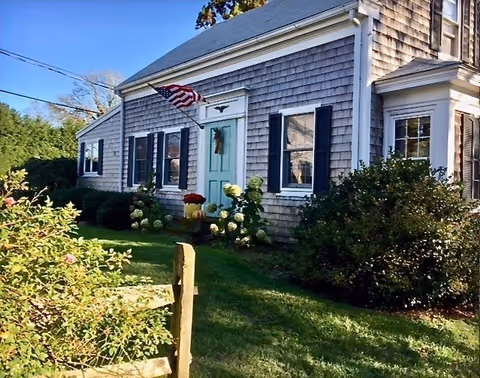Exterior front of a shingled house with a turquoise front door, American flag, and a green lawn with shrubs.