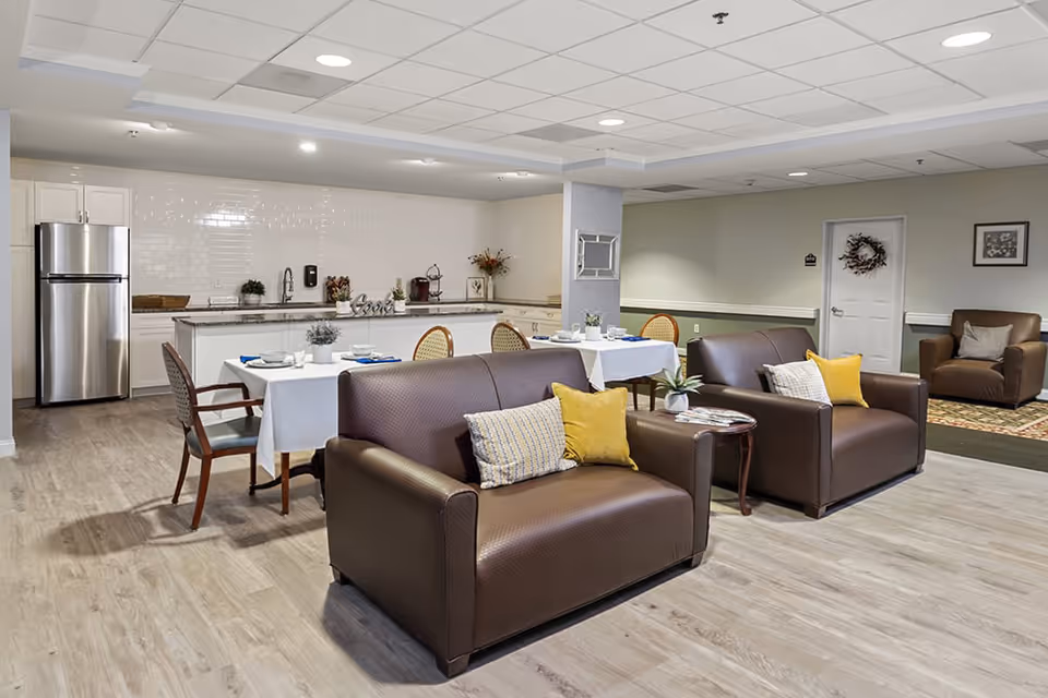 A spacious common area in a senior living facility featuring a kitchenette with a stainless steel refrigerator, white cabinets, and a tiled backsplash. In front of the kitchenette are two dining tables set with plates, cups, and napkins, surrounded by chairs. The seating area includes two brown leather loveseats with yellow and patterned throw pillows, a small wooden side table with a plant and magazines, and a brown armchair in the background. The room has light wood flooring and a drop ceiling with recessed lighting.
