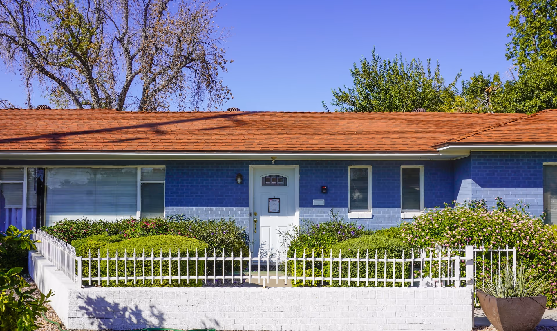 Front exterior view of a single-story assisted living home with blue brick walls, a white door, and a red shingle roof. The entrance is surrounded by neatly trimmed green bushes and a white metal fence. Trees and clear blue sky are visible in the background.