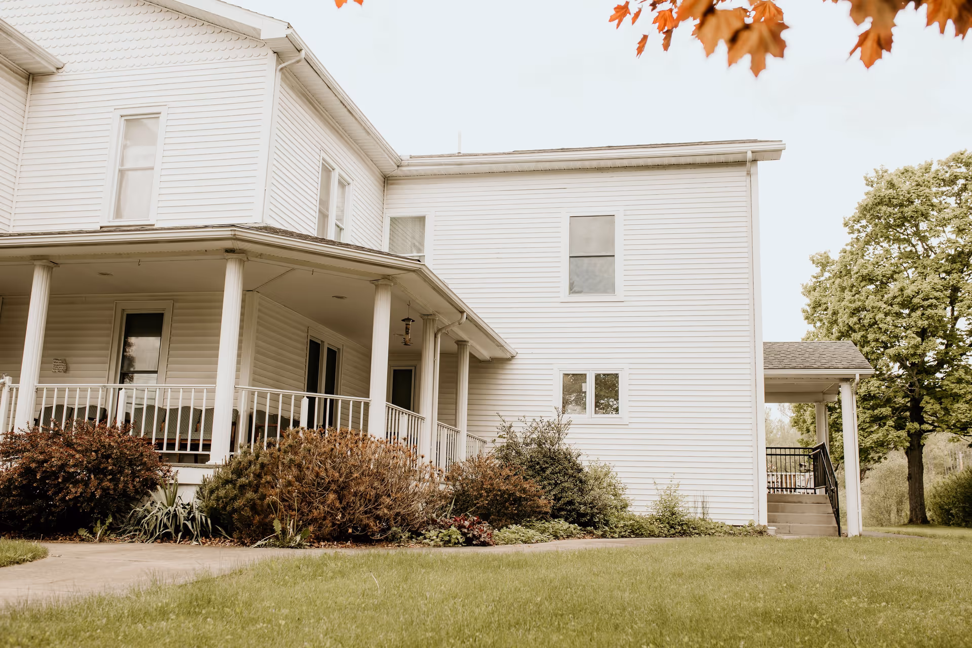 White two-story house with a covered wraparound porch, columns, shrubs, and a grassy lawn.