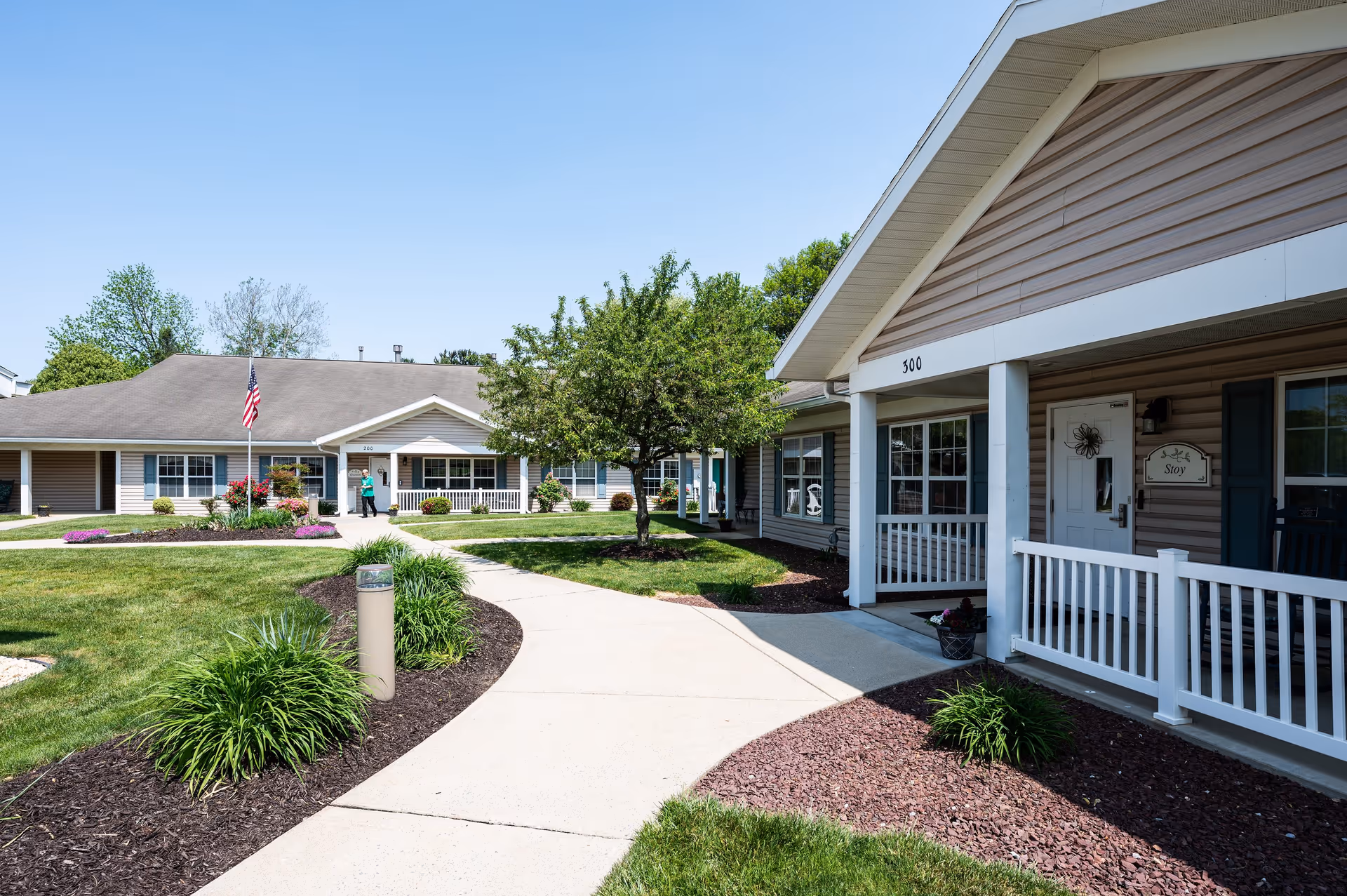 Exterior view of a senior living community building with beige siding, white trim, and a covered porch. A concrete walkway curves through a well-maintained lawn with shrubs and a tree. An American flag is visible near the center building, and a person is standing near the entrance.