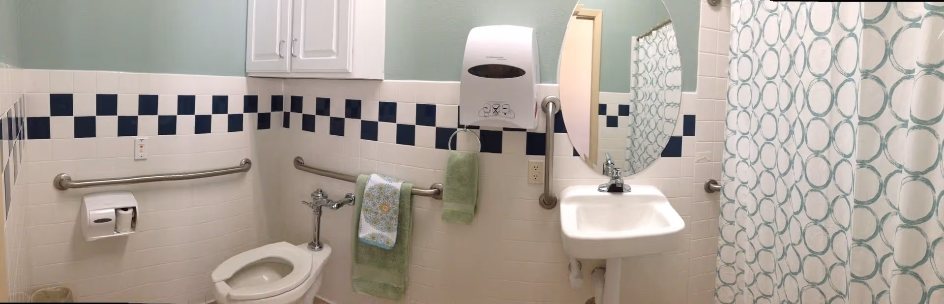 A clean and accessible bathroom with white tiled walls featuring a blue checkered border. The bathroom includes a toilet with grab bars on both sides, a white sink with a round mirror above it, a wall-mounted paper towel dispenser, and a shower area with a curtain decorated with blue circles. There are green and floral towels hanging on the grab bars.