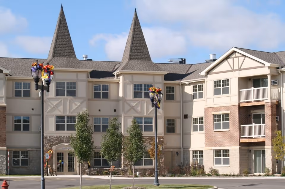 Exterior view of a multi-story senior living facility building with two pointed tower-like structures on the roof, multiple windows, balconies, and decorative street lamps adorned with fall-themed decorations. There are small trees and a grassy area in front of the building under a partly cloudy sky.