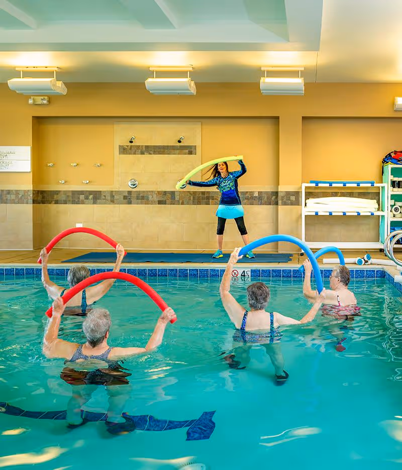An indoor swimming pool where four elderly women are participating in a water exercise class led by an instructor standing poolside holding a yellow pool noodle. The participants are holding red and blue pool noodles above the water. The room has beige tiled walls and shelves with towels and equipment.