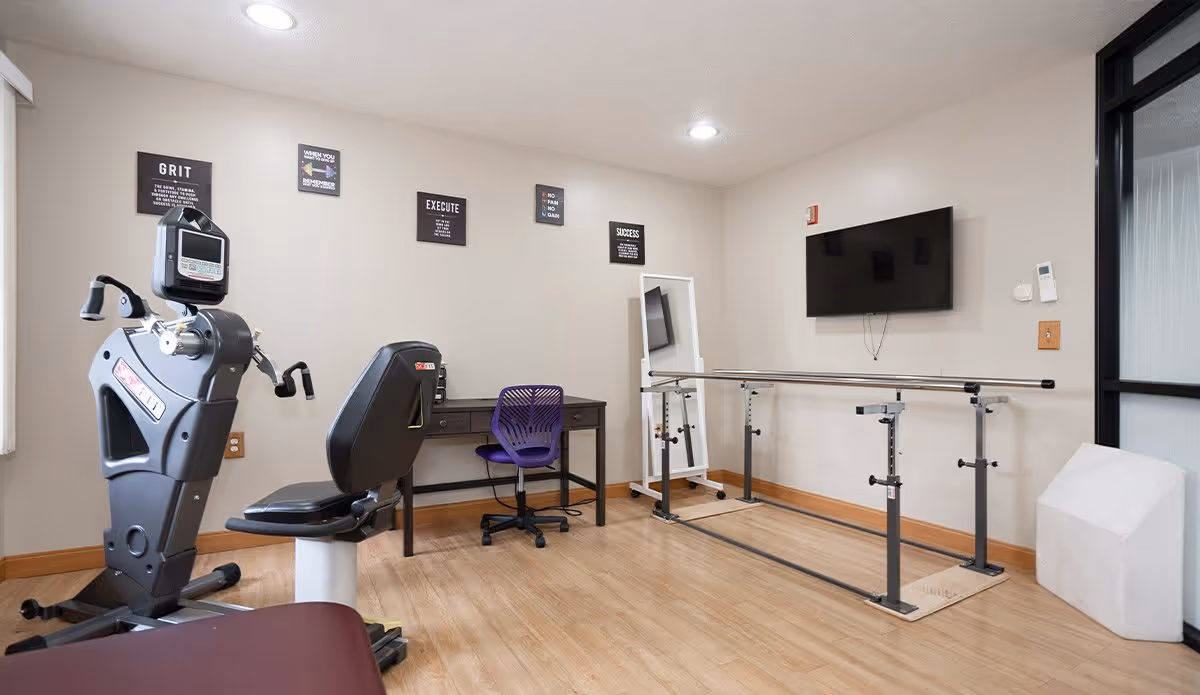 A small exercise room with a recumbent exercise bike, a purple office chair at a desk, a large mirror on a stand, parallel bars for walking support, a wall-mounted TV, and motivational signs on the wall.