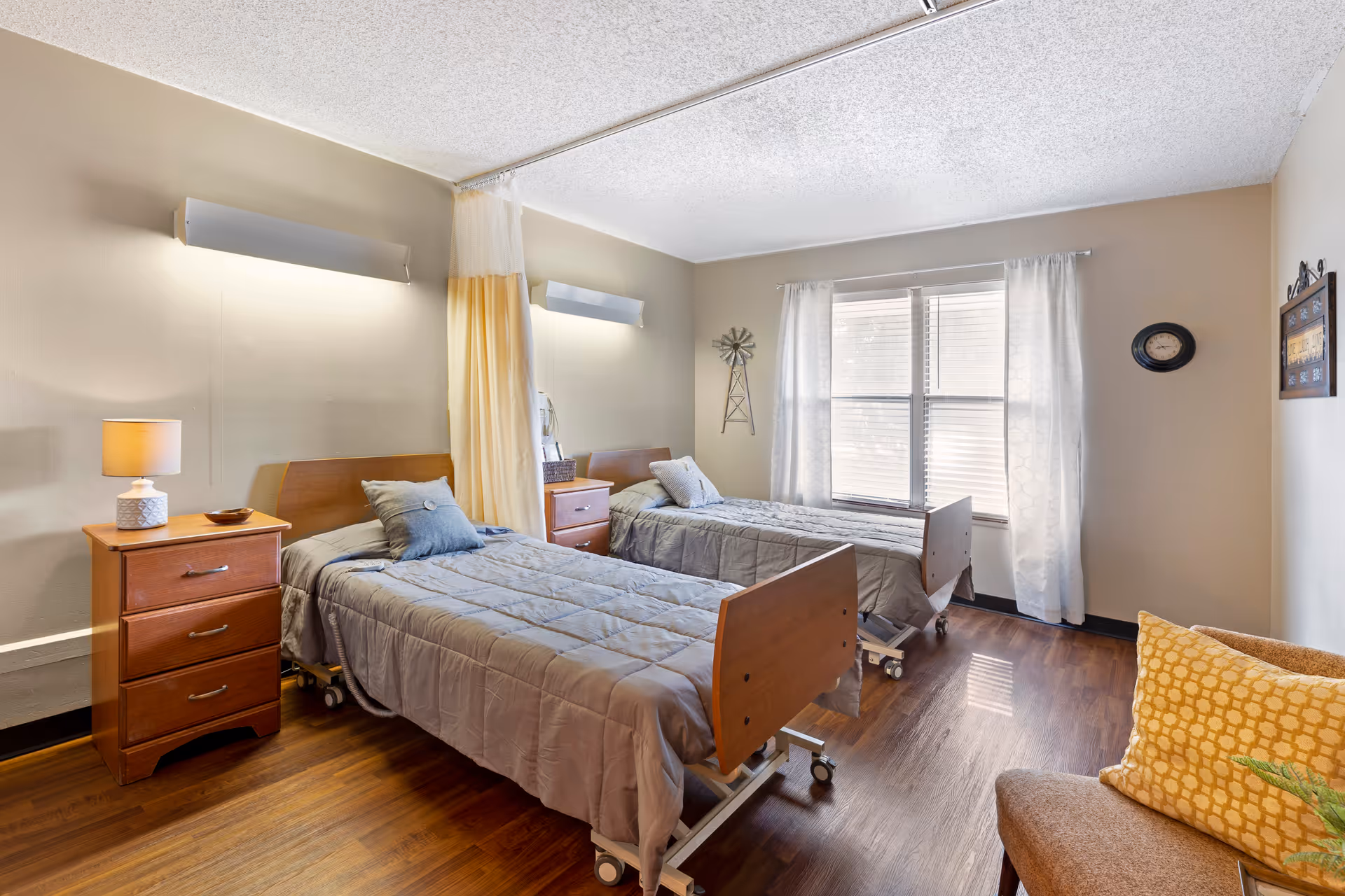 A bright and clean room with two hospital-style beds with gray bedding, separated by a curtain. Each bed has a wooden nightstand with a lamp and decorative items. The room has wooden flooring, a window with white curtains, a wall clock, and a small chair with a yellow cushion.