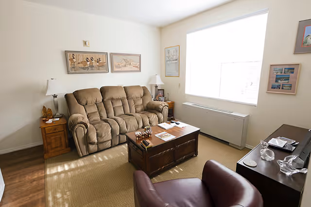 Furnished living room with a beige reclining sofa, coffee table, armchair, side tables, and a large window.