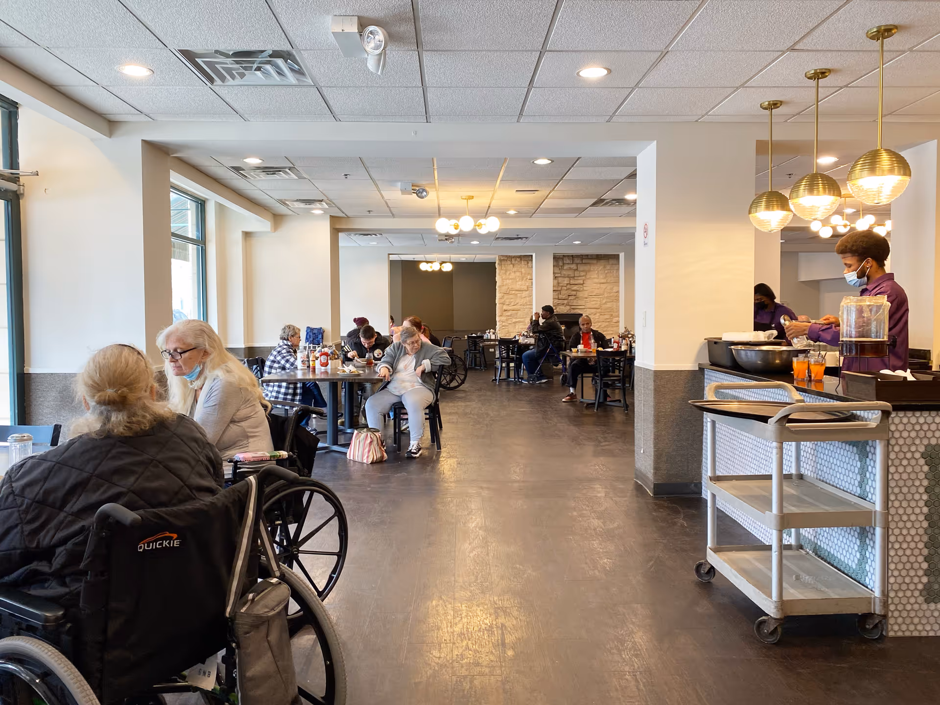 Interior view of a dining area in a senior living facility with several elderly residents seated at tables, some in wheelchairs, eating and socializing. A staff member wearing a mask is serving drinks from a cart near the right side of the image. The room has large windows on the left, modern lighting fixtures, and a clean, spacious layout.