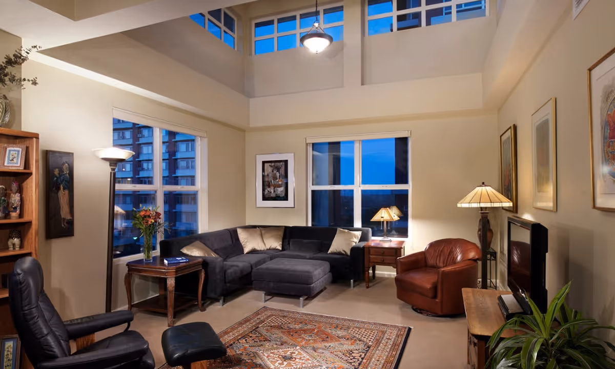 A cozy living room with a high ceiling and large windows showing a cityscape at dusk. The room features a dark gray sectional sofa with beige cushions, a brown leather armchair, a black recliner with matching ottoman, wooden side tables with lamps, framed artwork on the walls, a patterned area rug, and a flat-screen TV on a wooden stand. There is also a tall floor lamp and a bookshelf with decorative items.