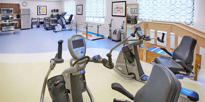 A senior living facility exercise room with various fitness equipment including recumbent bikes and arm exercise machines. The room has light-colored flooring, large windows with patterned curtains, framed artwork on the walls, and a small kitchenette area with a microwave, oven, and refrigerator in the background.
