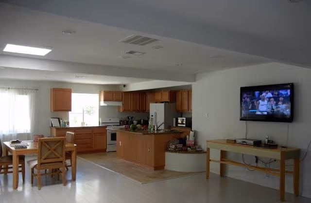 A bright, open interior space featuring a kitchen with wooden cabinets, a central island, and modern appliances. To the left, there is a dining area with a wooden table and chairs near a window with sheer curtains. On the right wall, a flat-screen TV is mounted above a wooden console table with electronic devices and cables underneath. The floor is light-colored, and the ceiling has recessed lighting and a skylight.