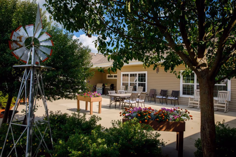 Sunlit courtyard patio with potted flowers, outdoor seating, and a decorative windmill in front of the assisted living building.