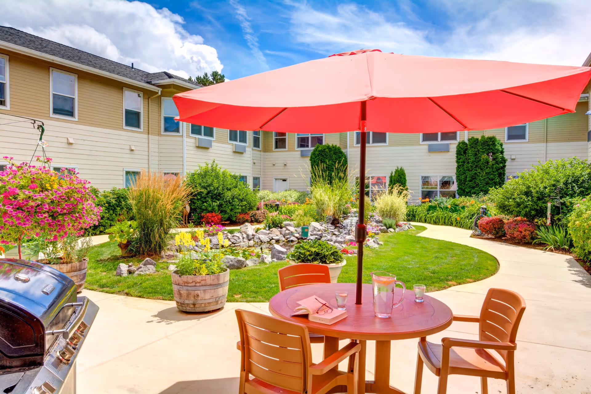 Outdoor courtyard area with a round table and four chairs under a large red umbrella. The table has a pitcher of water, two glasses, and an open book with reading glasses on it. Surrounding the courtyard are well-maintained garden beds with various plants and flowers, and a paved walkway curves through the garden. The building exterior is visible in the background under a partly cloudy blue sky.