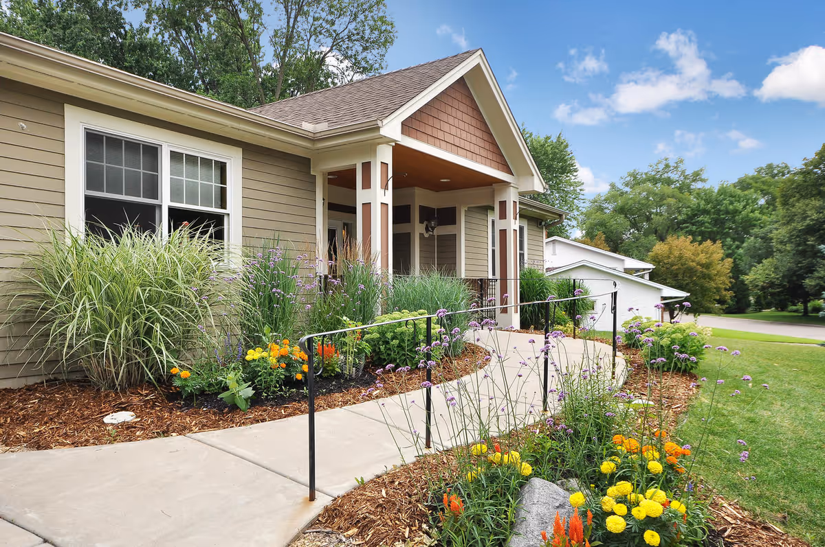 Exterior view of a single-story building with beige siding and white trim, featuring a covered entrance with a gabled roof. A concrete walkway with a black handrail curves through a landscaped garden with colorful flowers and green shrubs. Trees and a blue sky with some clouds are visible in the background.