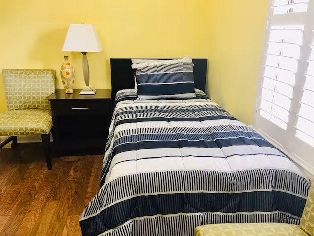 A small bedroom with a single bed covered in a striped blue, white, and black comforter and matching pillow. Next to the bed is a dark wooden nightstand with a white lamp and decorative vase. There is a green patterned chair to the left of the nightstand and another similar chair partially visible in the foreground. The walls are painted yellow and there is a window with white shutters on the right side.