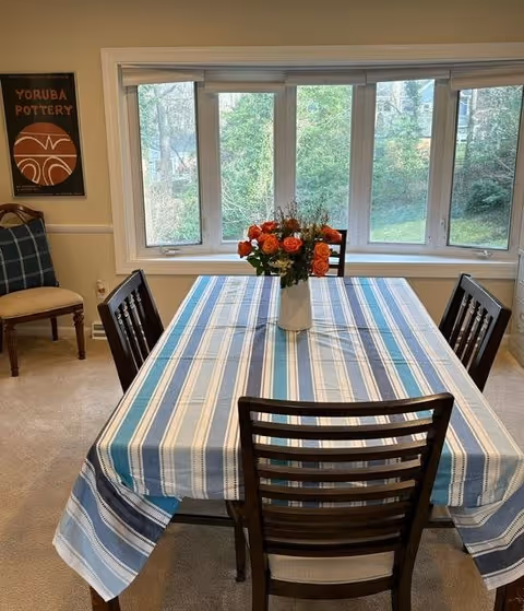 A dining room with a rectangular table covered by a blue and white striped tablecloth. Four dark wooden chairs surround the table. A vase with orange and red flowers is placed in the center of the table. Behind the table is a large window showing a green outdoor view. A framed artwork titled 'Yoruba Pottery' hangs on the left wall, and a cushioned chair with a plaid pattern is positioned nearby.