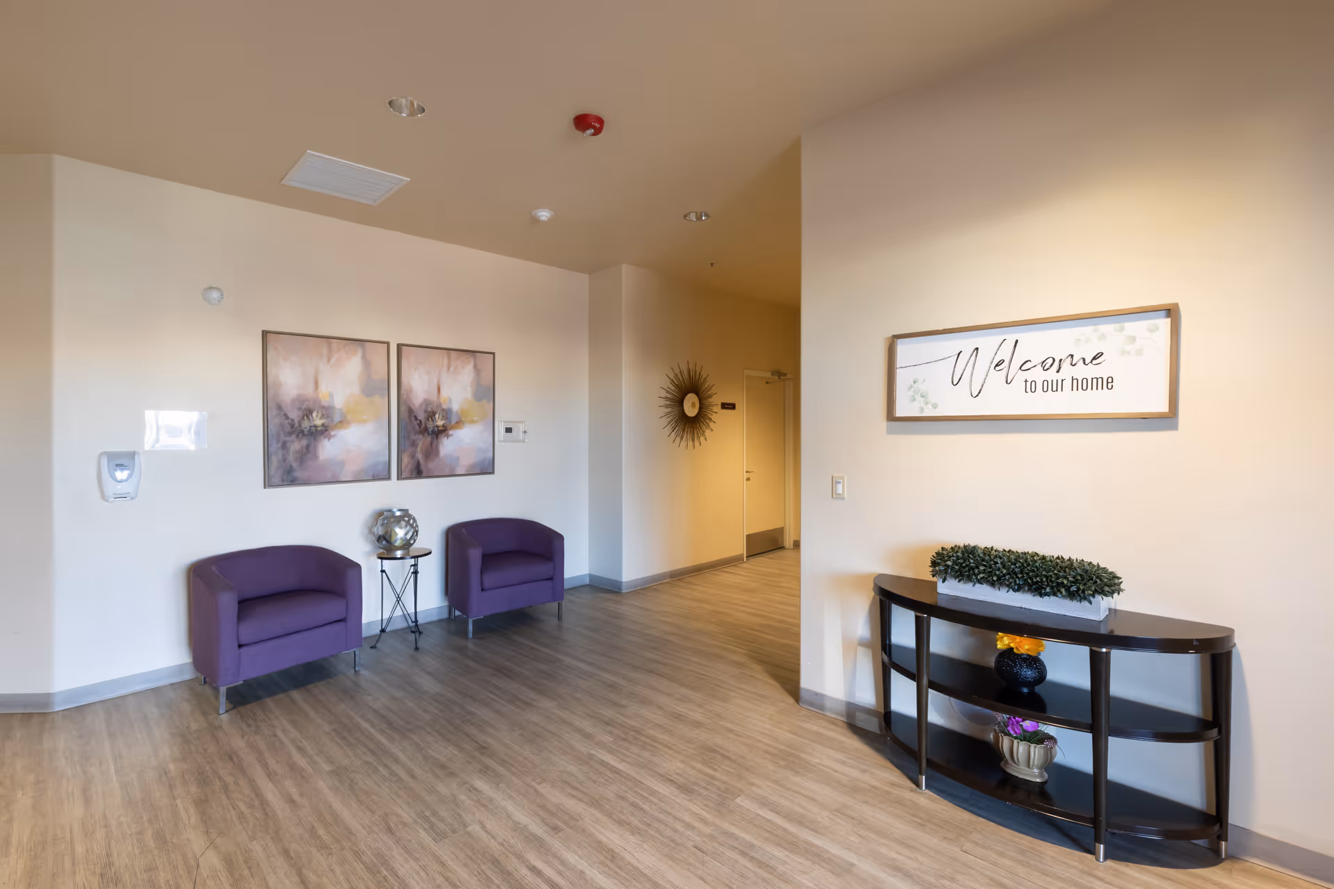 A welcoming interior hallway area with two purple armchairs and a small round table between them, decorated with a metallic globe. On the wall above the chairs are two abstract paintings. Across the hallway is a black console table with decorative plants and a framed sign above it that reads 'Welcome to our home'. The floor is wood-style laminate, and the walls are painted a light beige color.