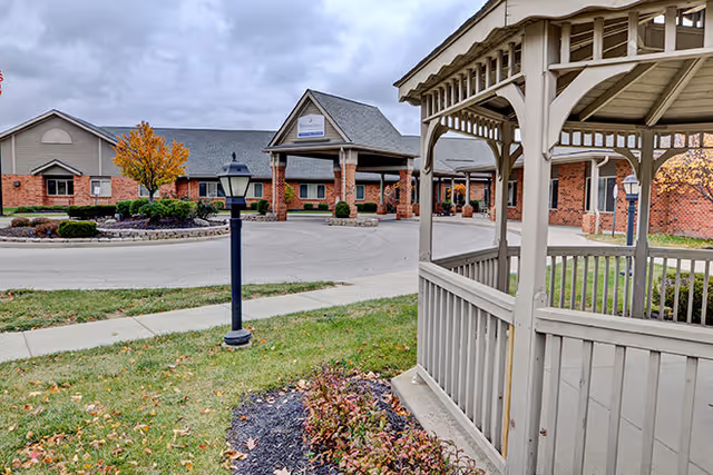 Exterior view of Brookdale Pinnacle facility showing a brick building with a covered entrance, a lamp post, a gazebo in the foreground, and some autumn trees and landscaping under a cloudy sky.