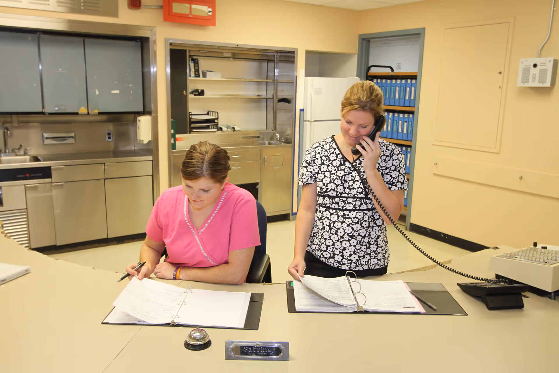 Two women working at a reception desk in an office-like environment. One woman in a pink scrub top is seated and writing in a binder, while the other woman in a black and white floral top is standing and talking on a corded telephone, also looking at a binder. The background shows stainless steel cabinets, a refrigerator, and shelves with blue binders.