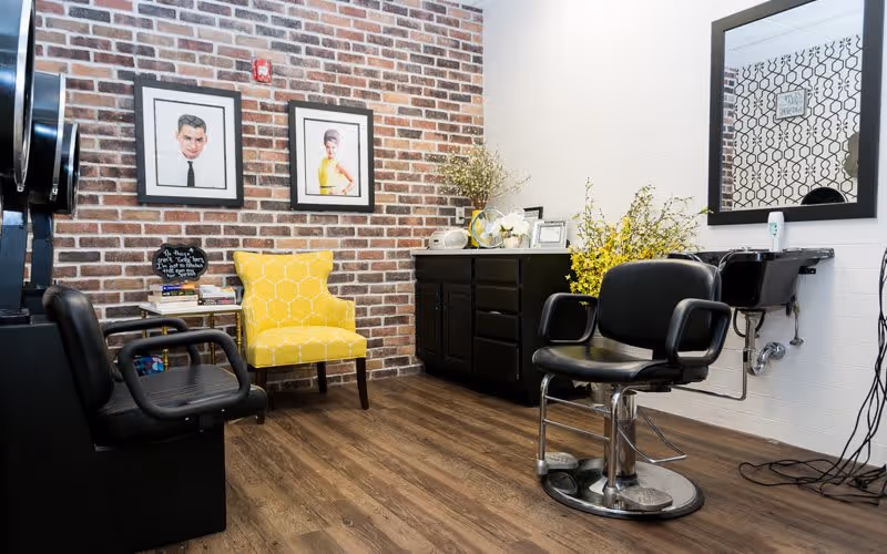 Interior of a salon area with a black salon chair in front of a large mirror on the right, a black cabinet with decorative plants and framed pictures on top, a yellow patterned armchair against a brick wall, and two framed portraits hanging on the brick wall.