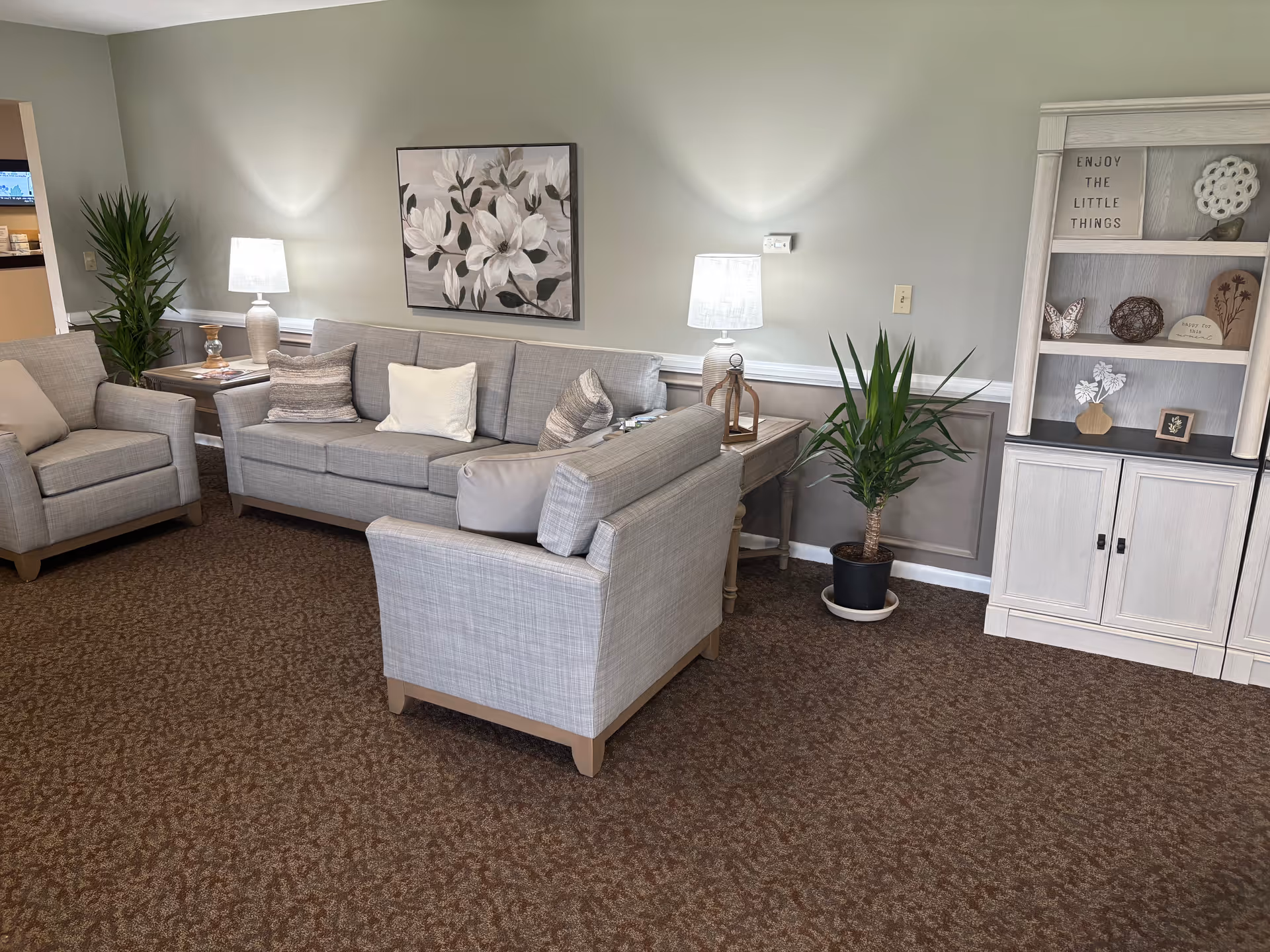 A cozy living room with gray sofas and armchairs, side tables with lamps, potted plants, wall art, and a white display cabinet with decorations.