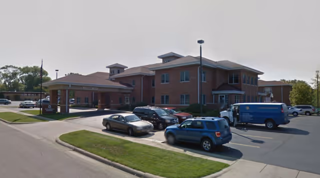 Exterior view of a two-story brick senior living facility with a covered entrance and several parked cars in the parking lot under a clear sky.