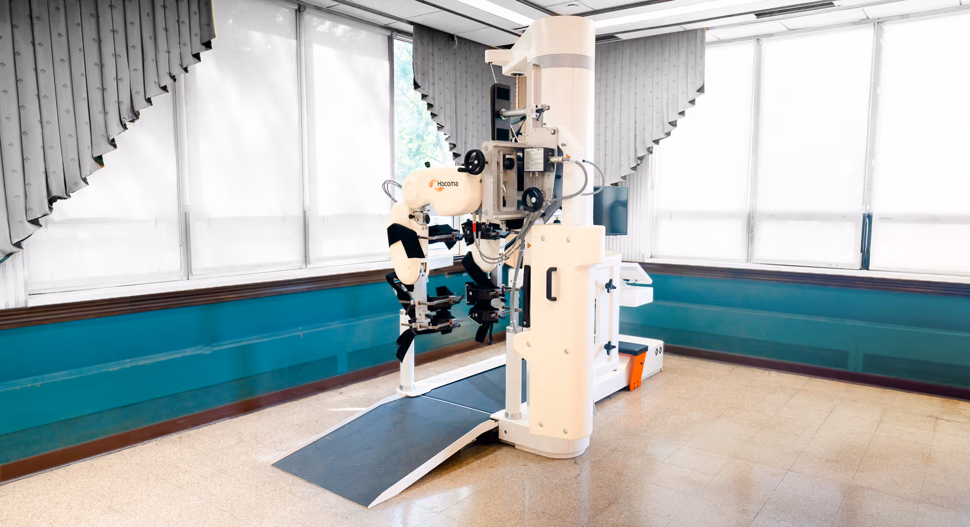 A medical rehabilitation robot device in a bright room with large windows covered by white blinds and gray curtains. The device is designed for physical therapy, featuring mechanical arms and supports for assisting leg movement. The room has teal walls with a wooden trim and a beige tiled floor.