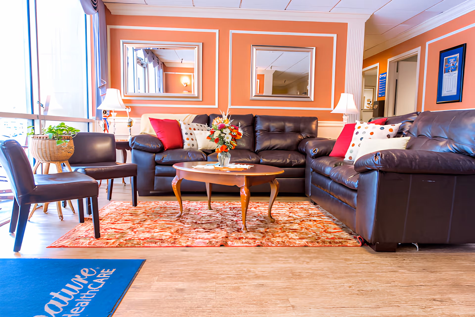 Bright nursing home sitting area with leather sofas, armchairs, a coffee table with flowers and a patterned rug.