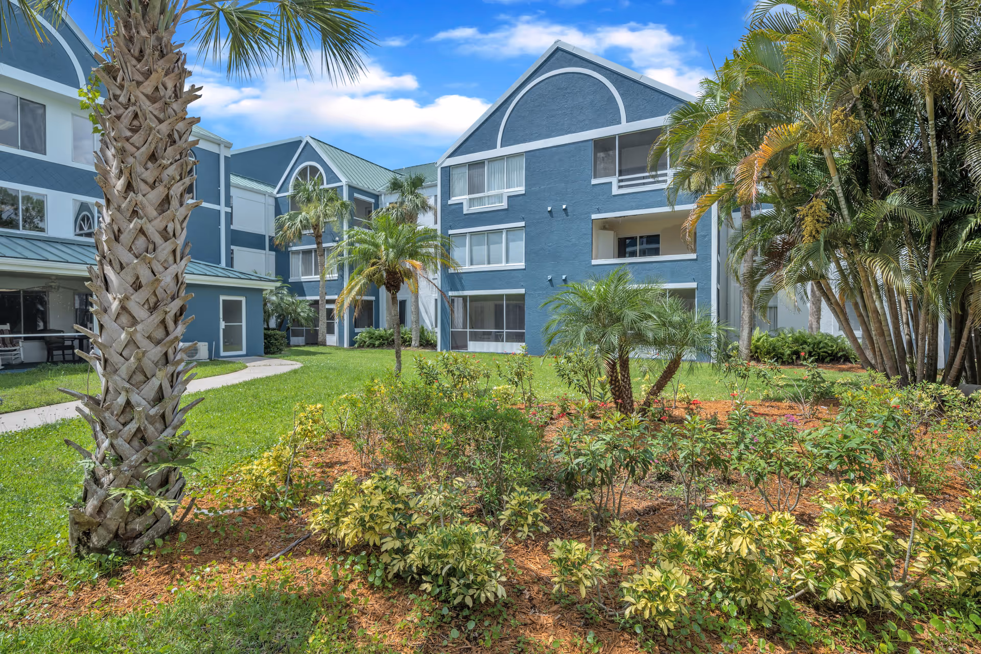 Exterior view of a senior living facility with blue buildings featuring multiple windows and balconies, surrounded by green lawns, palm trees, and landscaped garden beds under a partly cloudy blue sky.