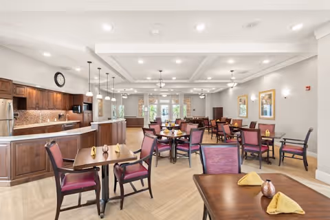 A spacious dining room in a memory care community featuring multiple wooden tables with purple cushioned chairs. Each table is set with folded yellow napkins and small decorative items. The room has a modern kitchen area with wooden cabinets, a clock on the wall, and pendant lighting. The walls are light-colored with framed artwork, and large windows at the far end let in natural light.