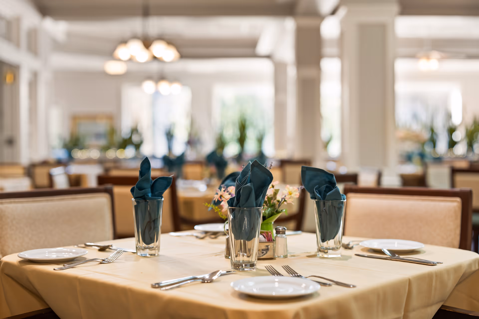 A dining table set for four in a well-lit dining room with beige cushioned chairs, white plates, silverware, glasses with folded dark green napkins, and a small floral centerpiece. The background shows more tables and chairs in a spacious, elegant dining area with soft lighting.