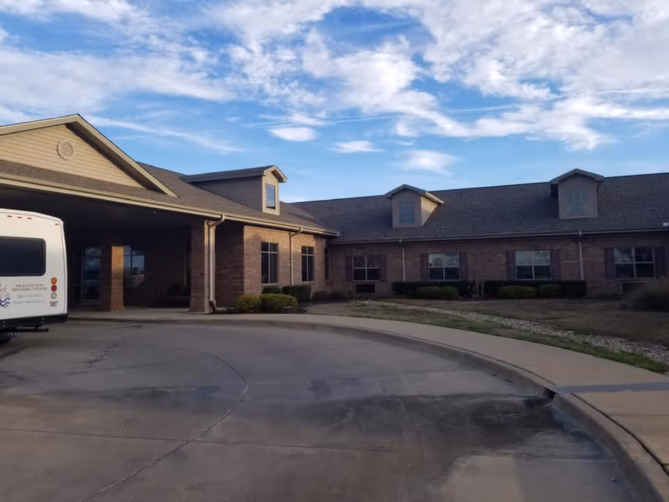 Exterior view of a single-story brick building with multiple windows and dormer windows on the roof under a partly cloudy sky. A curved driveway leads to the entrance, and a white van is parked on the left side near the building.