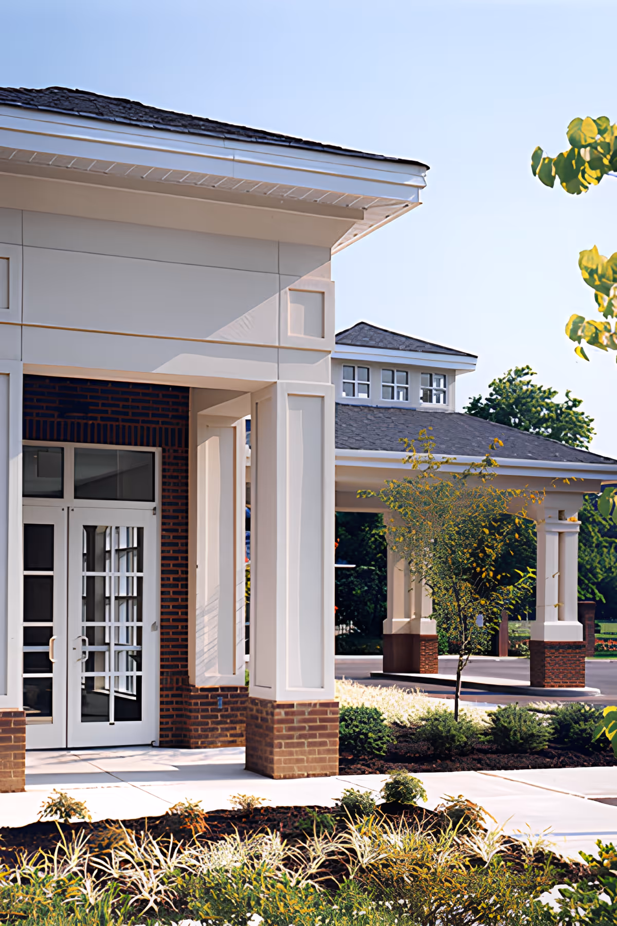 Entrance area of a building with white pillars and brick accents, featuring glass double doors and a covered driveway. There are landscaped plants and small trees around the walkway.