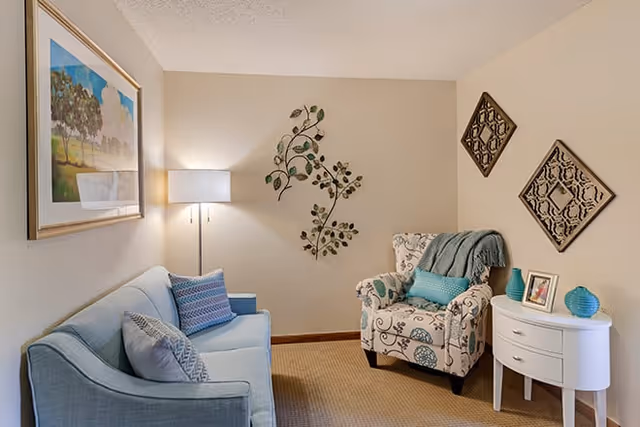 Cozy living room with a light blue sofa, patterned armchair, floor lamp, wall art, and a small white side table.