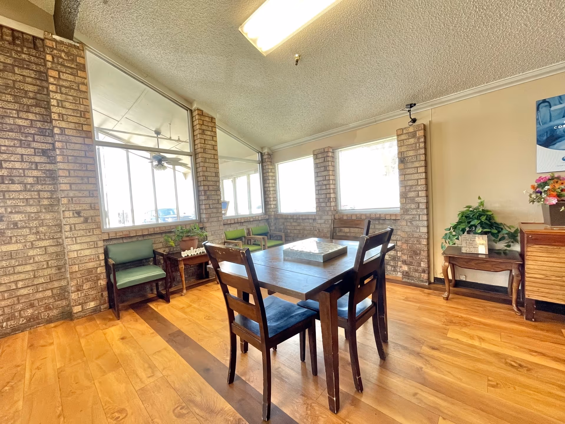 A bright interior room with wooden flooring and brick walls featuring large windows. There is a wooden table with four chairs in the center, two green cushioned chairs along the wall, and small tables with plants and decorative items. The room has a welcoming and cozy atmosphere.