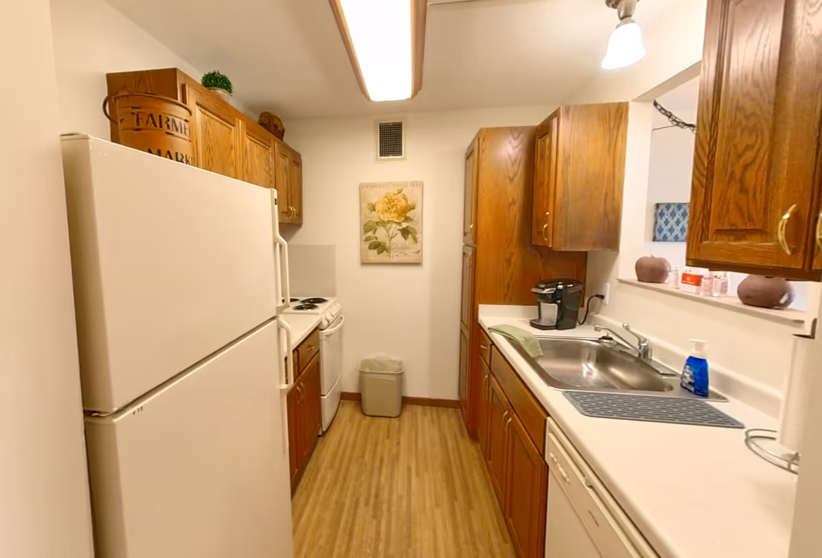 A narrow kitchen with wooden cabinets on both sides, a white refrigerator on the left, a white stove and oven next to it, and a stainless steel sink with a coffee maker on the right countertop. The floor has a wood-like finish, and there is a floral painting on the wall at the end of the kitchen.