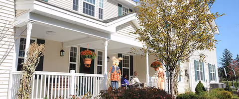 Front exterior view of a white multi-story residential building with a covered porch, white railing, hanging flower pots, and two scarecrow decorations near the entrance. A tree with autumn leaves is visible in the foreground under a clear blue sky.