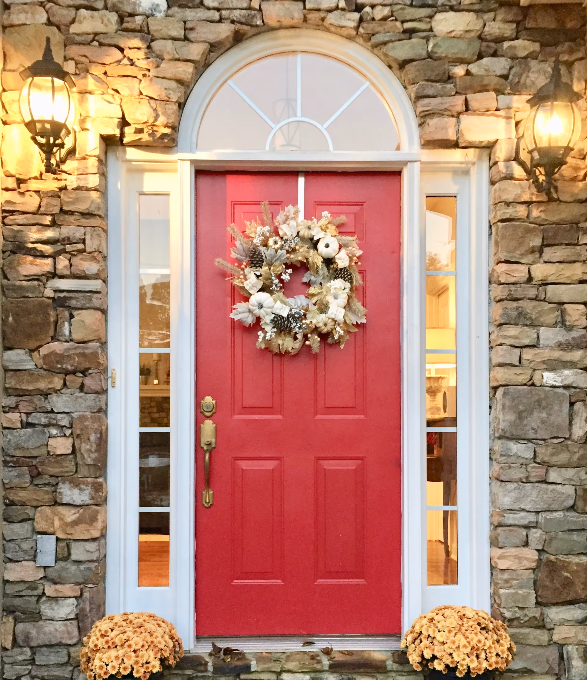 A red front door with a seasonal wreath set in a stone facade, flanked by glass sidelights and lanterns with potted mums on the step.