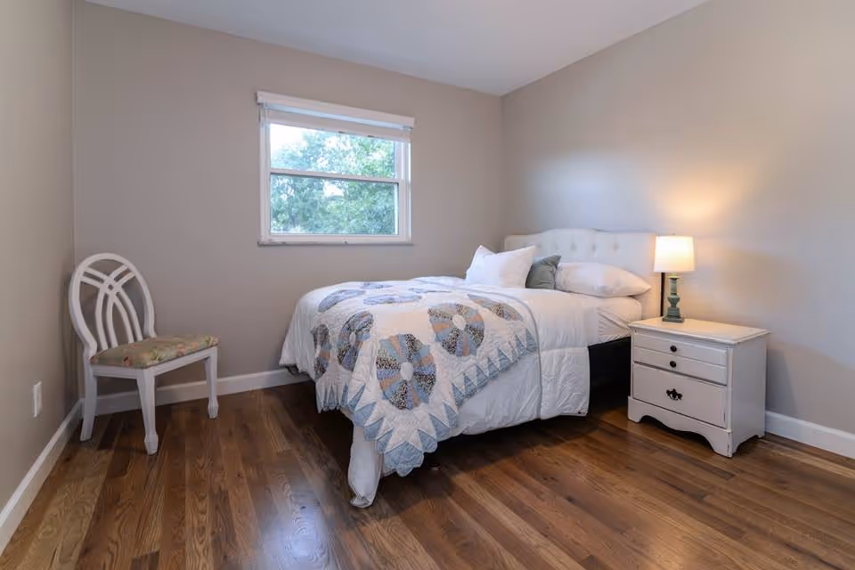 A simple bedroom with a bed covered in a white and patterned quilt, a white nightstand with a lamp, a white chair with a floral cushion, and a window showing green trees outside. The room has wooden flooring and beige walls.