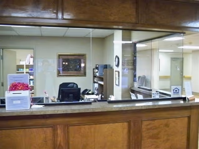 Reception desk area with a wooden counter and a glass partition. Behind the desk are office supplies, a computer, a framed picture on the wall, and an open door leading to another room.