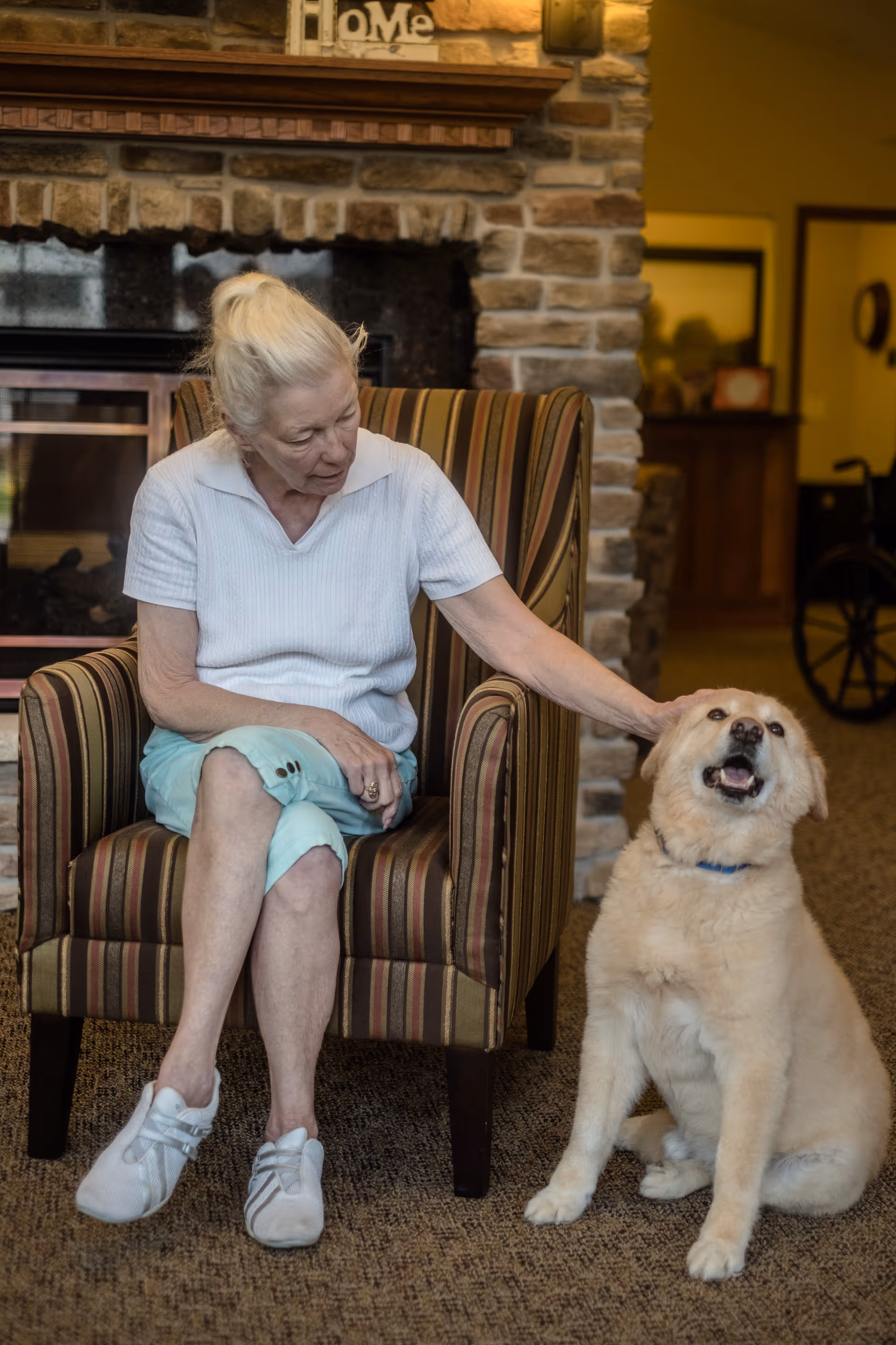 An elderly woman with white hair tied back sits in a striped armchair in a cozy room with a stone fireplace behind her. She is wearing a white short-sleeve shirt, light blue pants, and white sneakers. She is gently petting a light-colored dog sitting on the carpeted floor next to her, which looks happy with its mouth open.