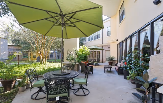 Outdoor patio area at Rosemont Assisted Living And Memory Care with a round metal table and four chairs with green cushions under a large green umbrella. The patio is surrounded by potted plants and greenery, with a building featuring large windows in the background.