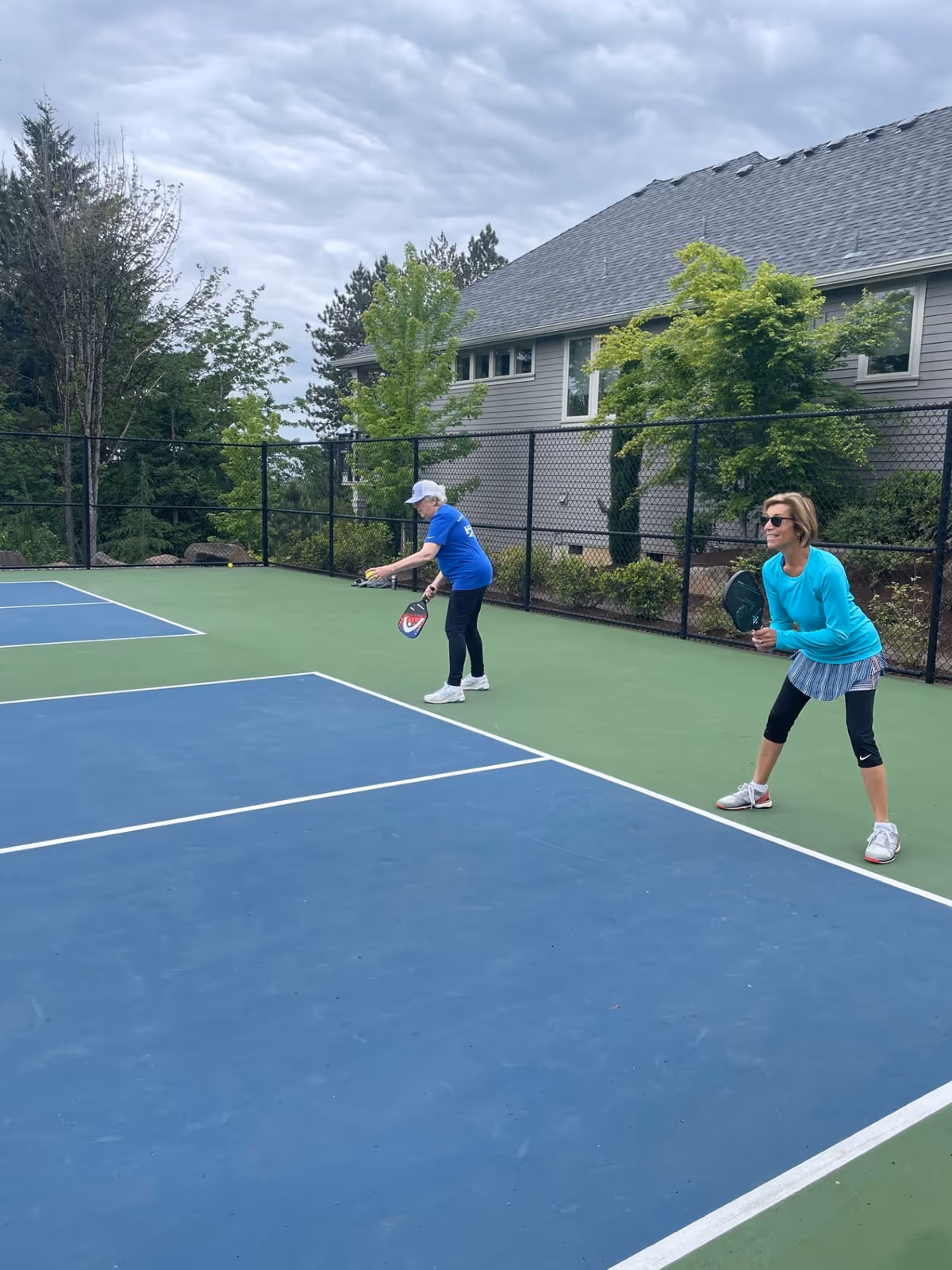 Two women playing pickleball on an outdoor court surrounded by a black chain-link fence, with trees and a building in the background under a cloudy sky.